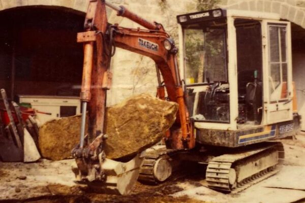 Old photo of orange takeuchi parked in front of building on a building site with operator door open