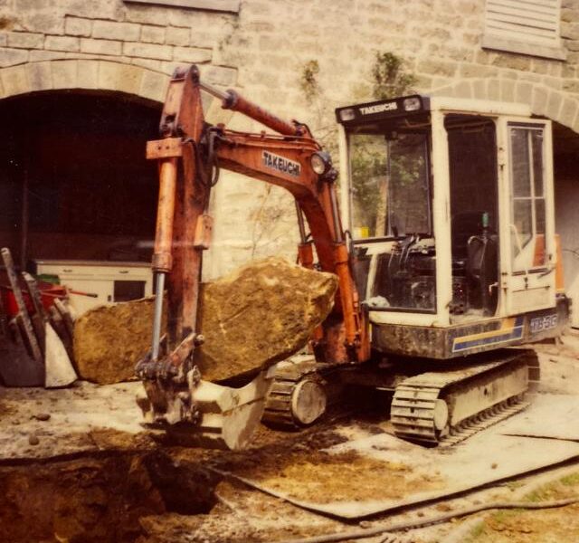 Old photo of orange takeuchi parked in front of building on a building site with operator door open