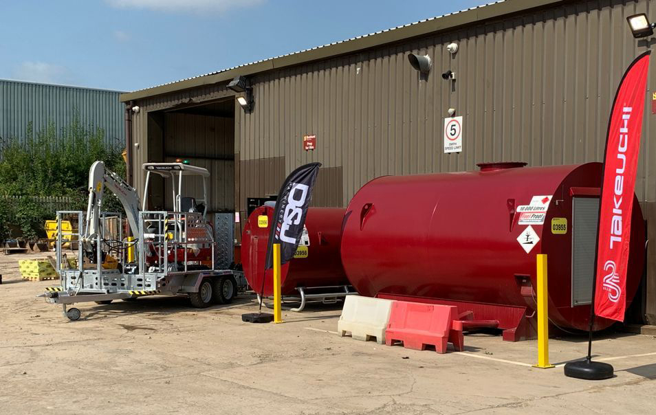 A digger on a trailer in front of a building with a takeuchi flag on the right hand side