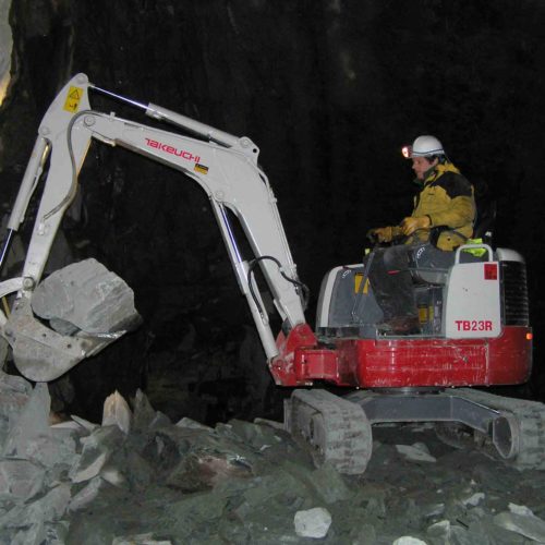 oldest working Takeuchi TB23R digger picking up a large boulder in the dark clearing a construction site